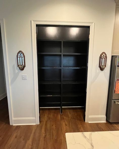 Open closet with black shelving in a white hallway, framed by decorative wall sconces.