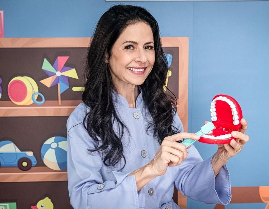 Mulher com cabelos escuros e uniforme azul demonstrando como escovar os dentes com uma modelo em uma sala de jogos colorida.