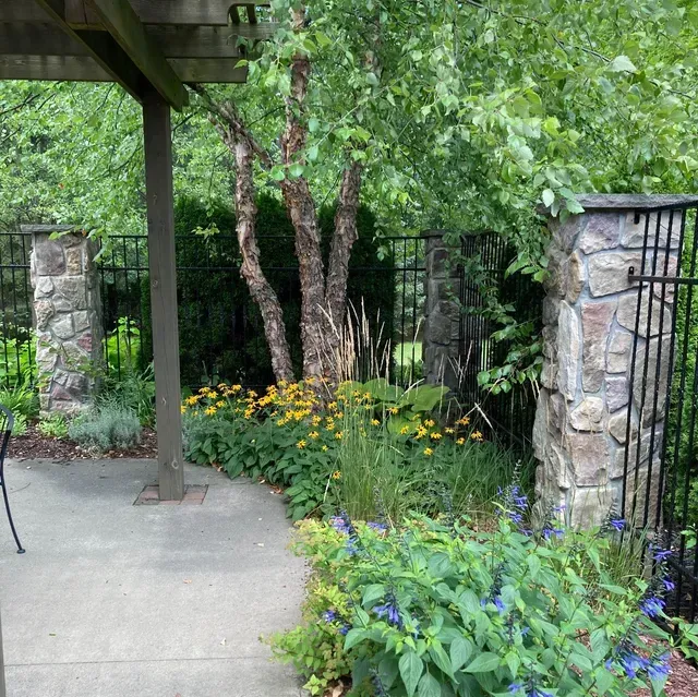 Patio with stone pillars, black fence, lush garden, birch tree, and yellow flowers.