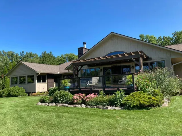 Beige house with a dark wooden deck and pergola, surrounded by a green lawn and garden, under a blue sky.