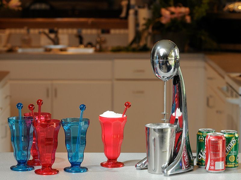 Milkshake machine with red and blue glasses, a red milkshake, and soda cans on a kitchen counter.