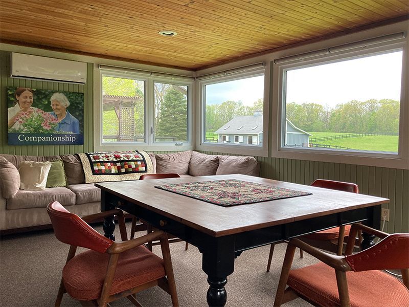 A light-filled sunroom with a large table and chairs. A sofa and artwork are on the left. Windows overlook a green landscape.