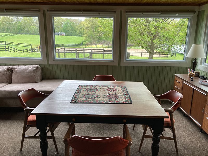 Dining room with a table and chairs, overlooking a field with horses and a tree through windows.
