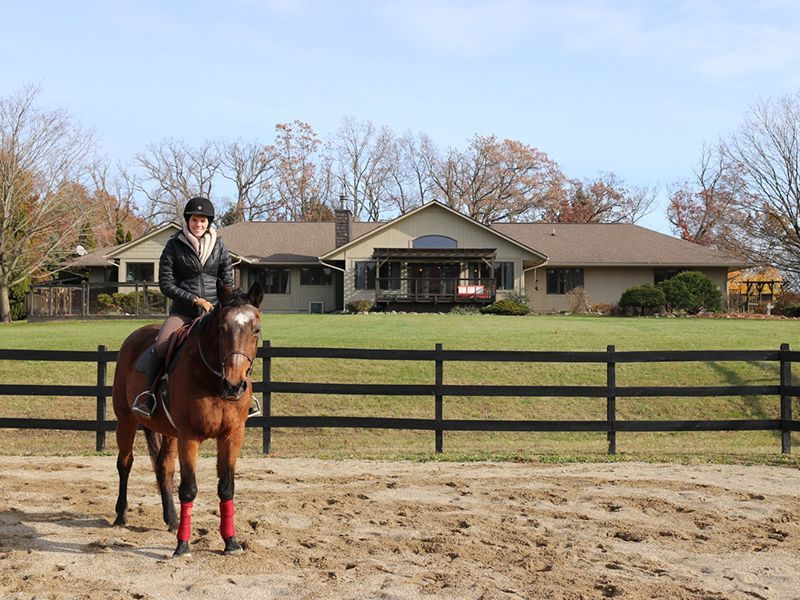 Woman riding a brown horse in front of a house. Black fence, sunny day.