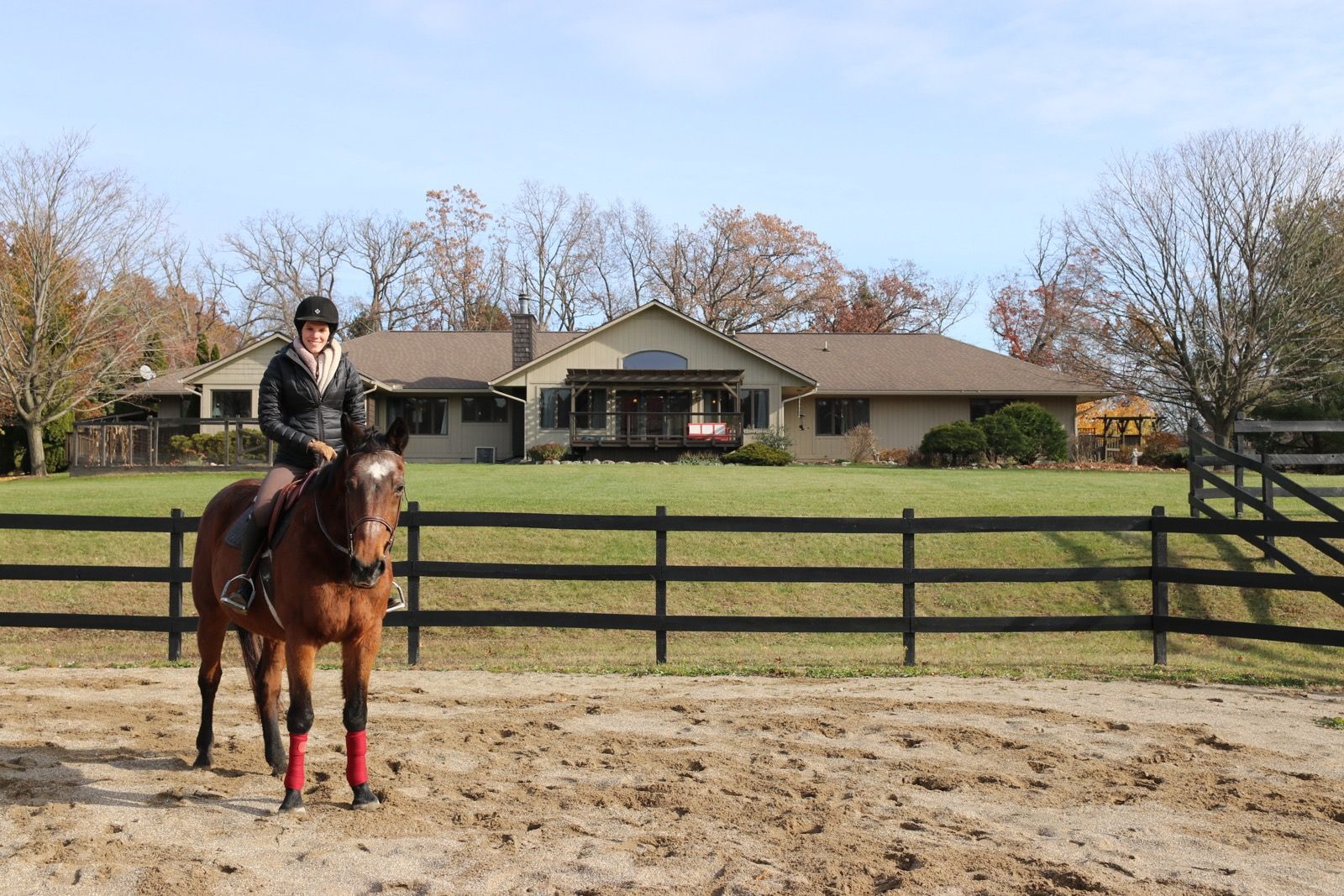 Person riding a brown horse in an arena, with a house and fence in the background under a blue sky.