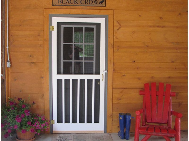 White screen door with black screen, vertical bars, and upper window, set in a wooden cabin wall.