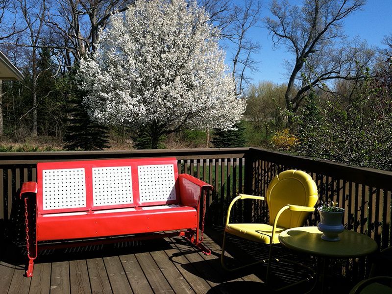 Red bench and yellow chair on a wooden deck, with a flowering tree in the background under a blue sky.