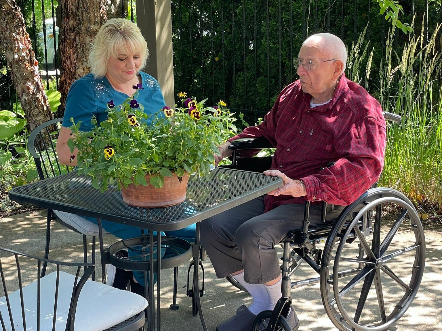 Woman and man in wheelchair at outdoor table with potted flowers.