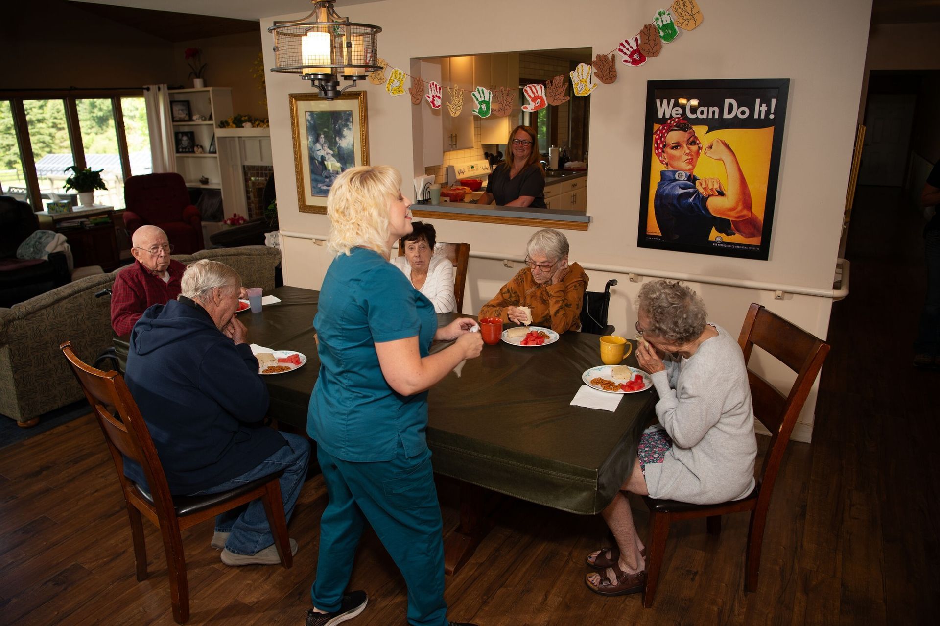 Caregiver serving food to seated people at a dining table.