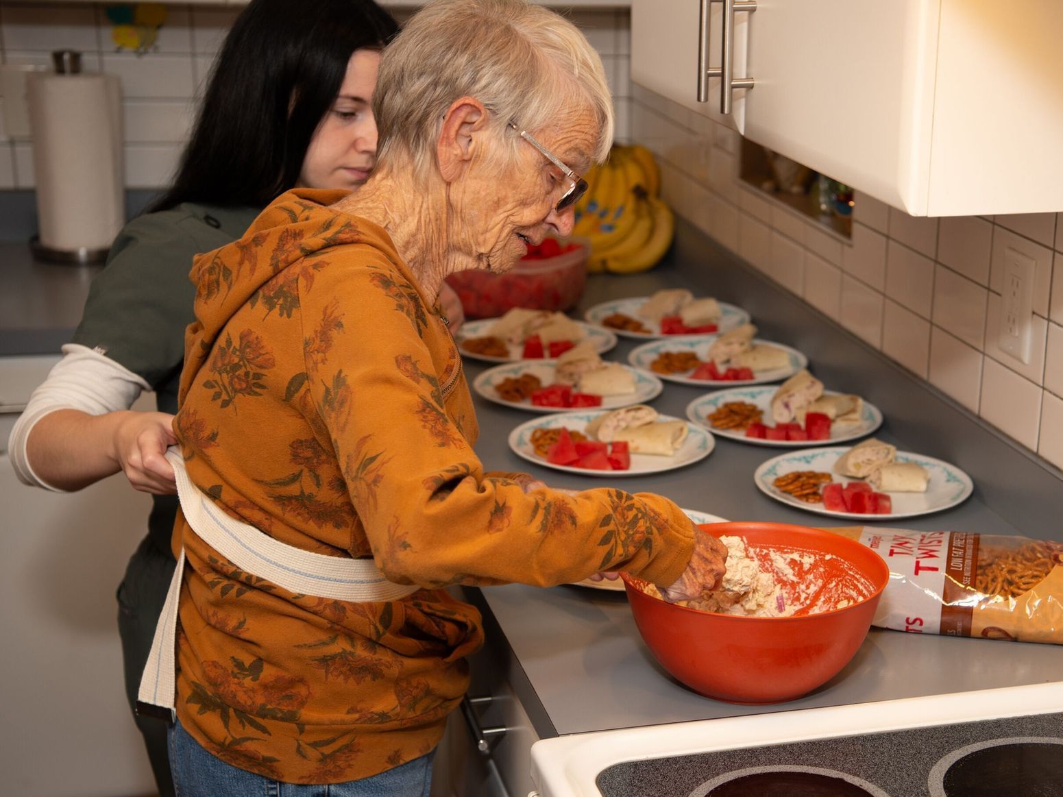 Woman assisted by another in a kitchen preparing food, plates laid out, orange bowl and brown bread visible.