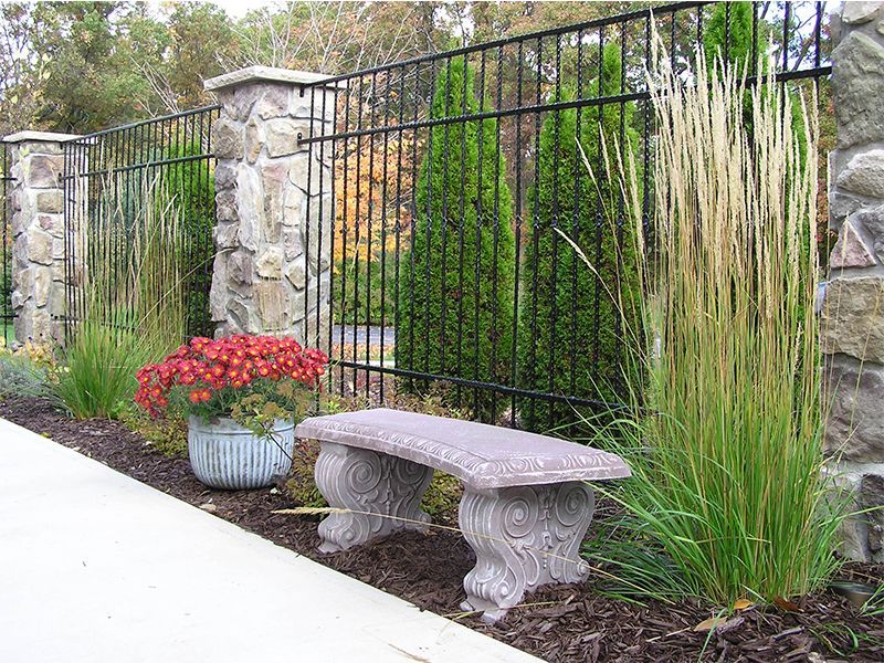 Stone bench in front of a black metal fence with stone pillars. Red flowers, green shrubs, and tall grass in the garden.
