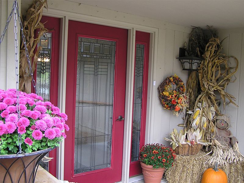 Red front door with fall decorations: wreath, scarecrow, mums, and pumpkins.