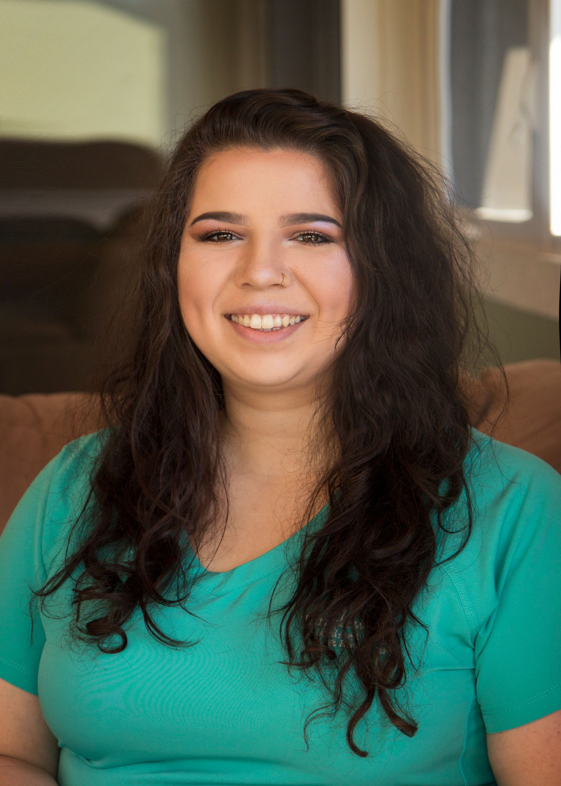 Woman with dark curly hair smiling, wearing a teal v-neck shirt, sitting indoors.