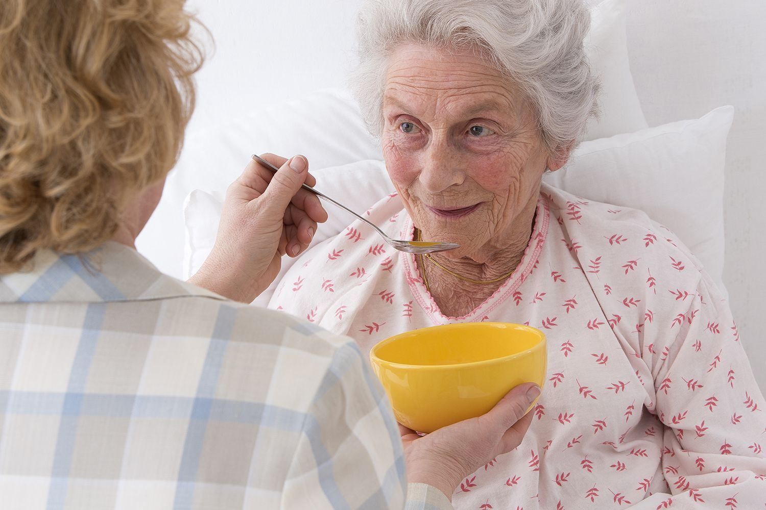 Woman in bed being fed by another person from a yellow bowl.