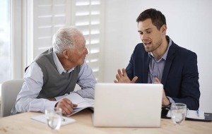 One older man and one younger man sitting together at a table looking at a laptop.
