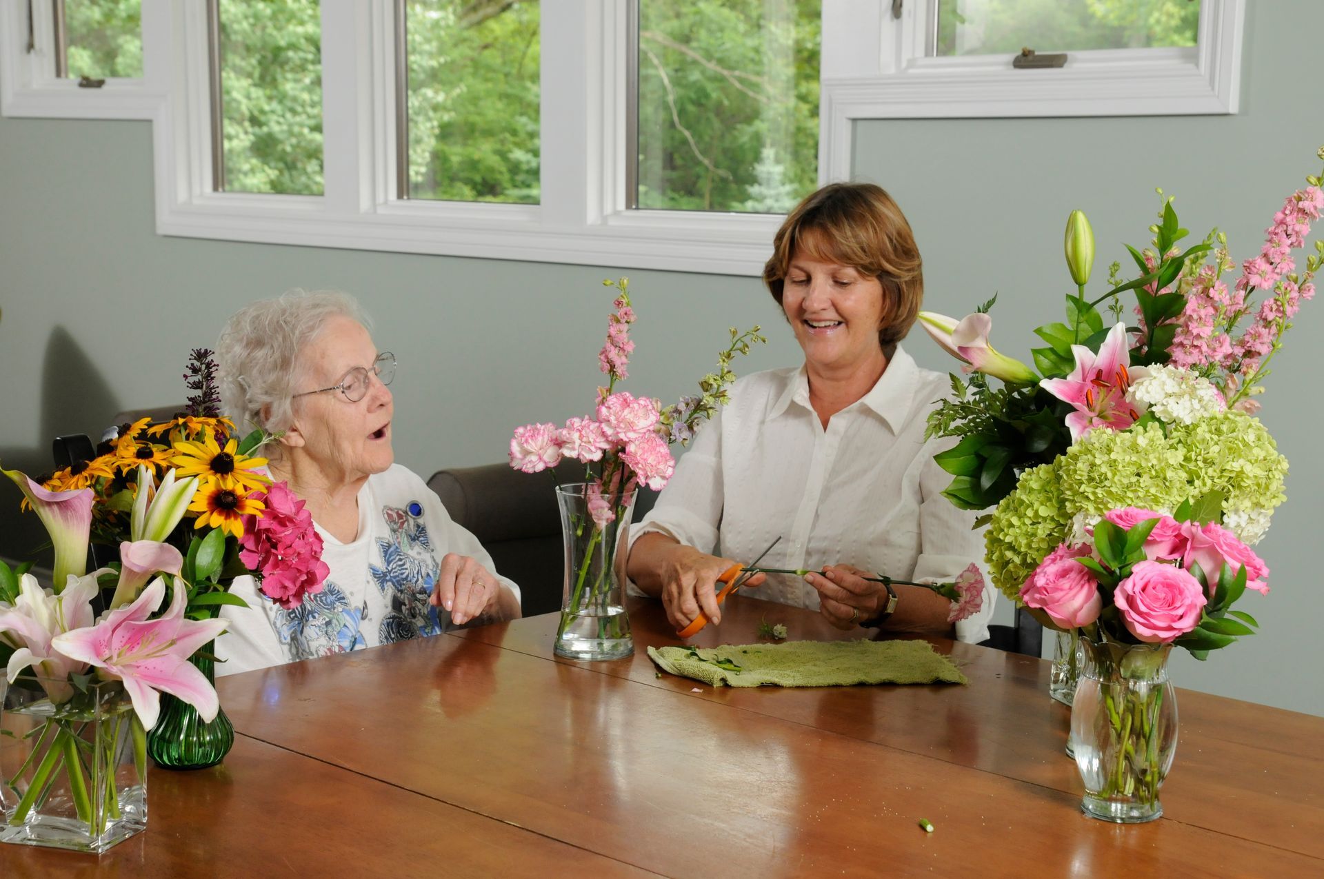 Woman and older woman arranging flowers at a table, smiling. Bright floral bouquets and greenery are visible.