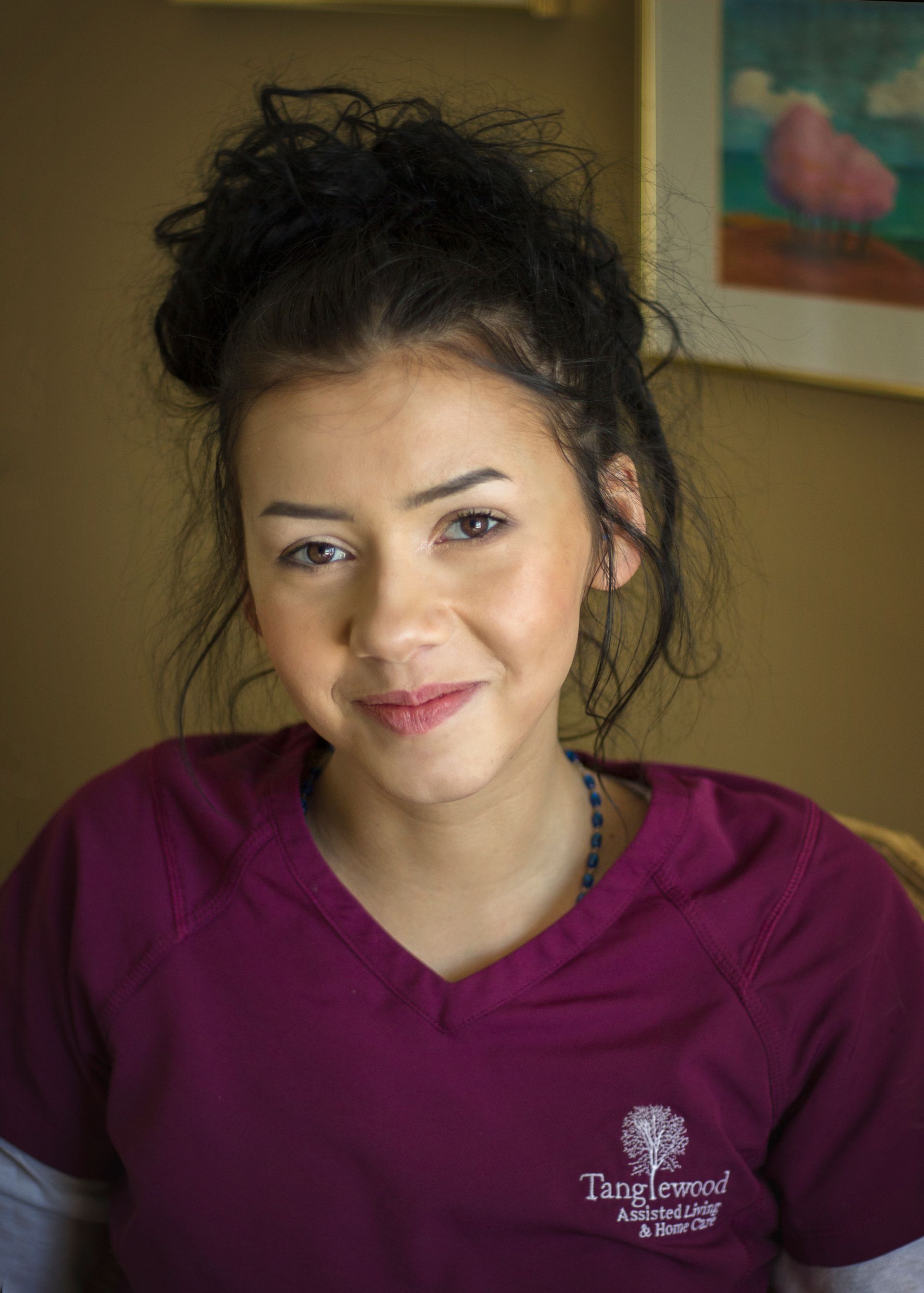 Woman wearing a maroon uniform smiles, hair up, sitting indoors. A painting is in the background.
