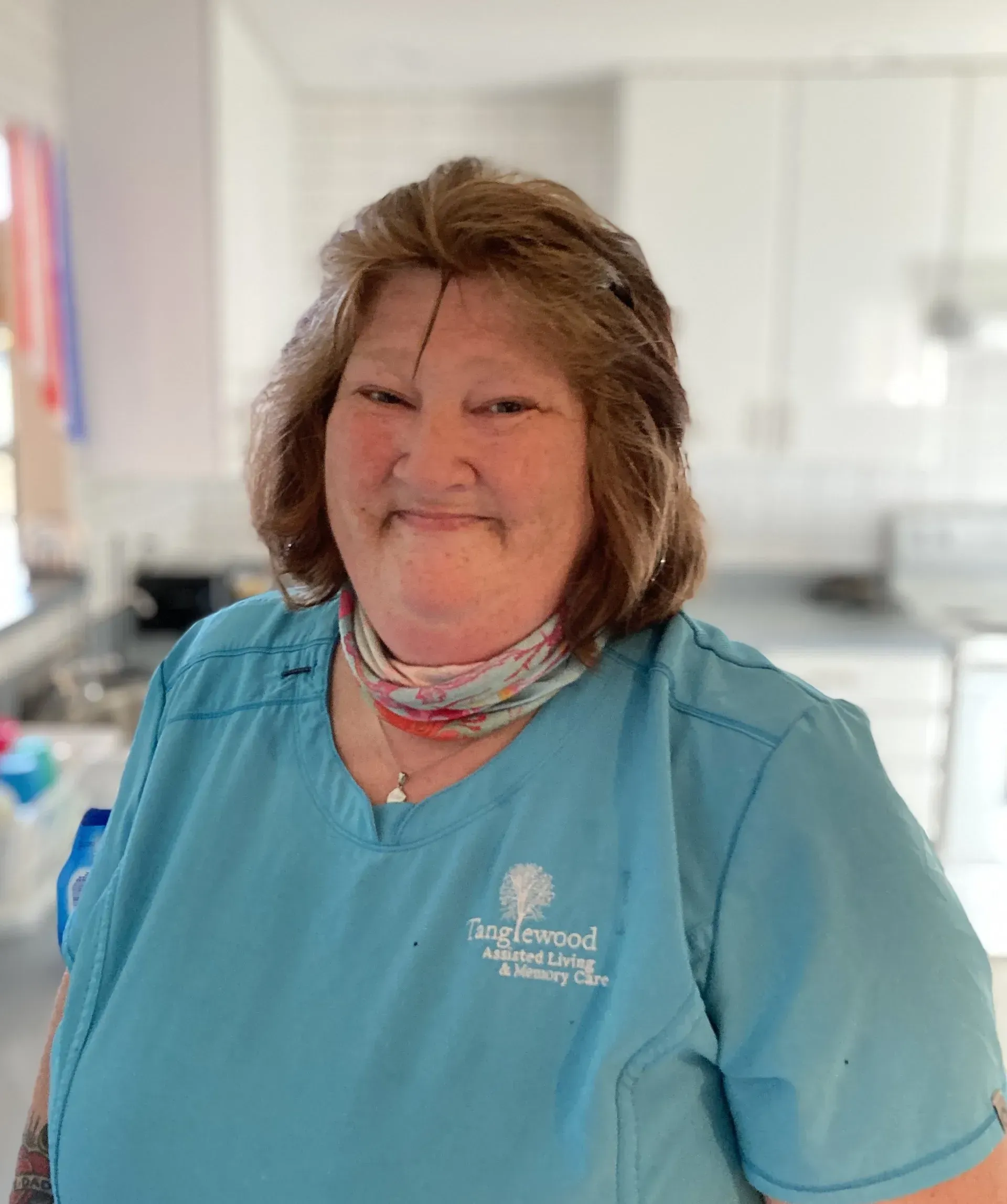 Woman in blue scrubs, smiling, with a neck scarf, in a kitchen setting.