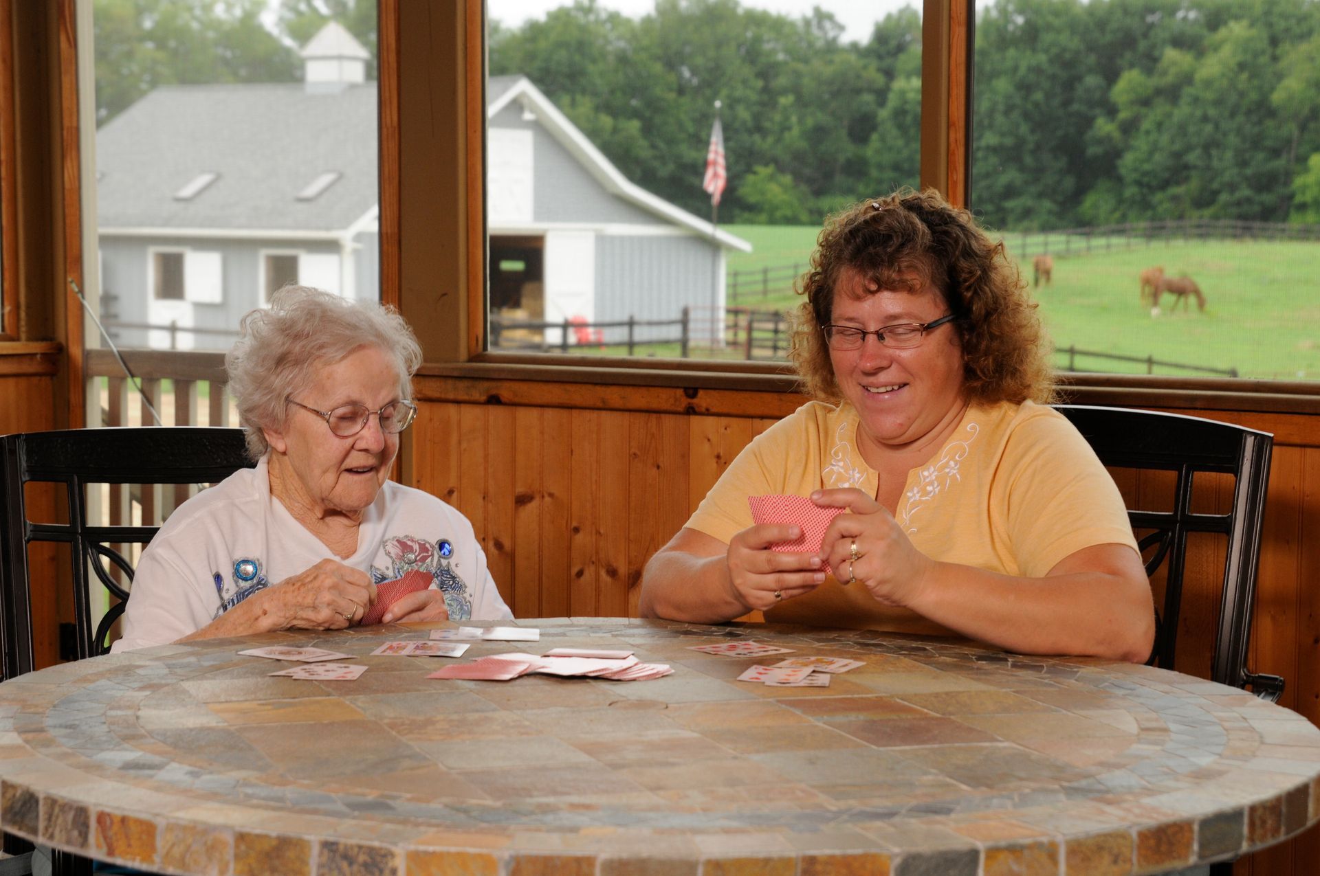 Two women playing cards at a round table, barn and horses visible in the background.
