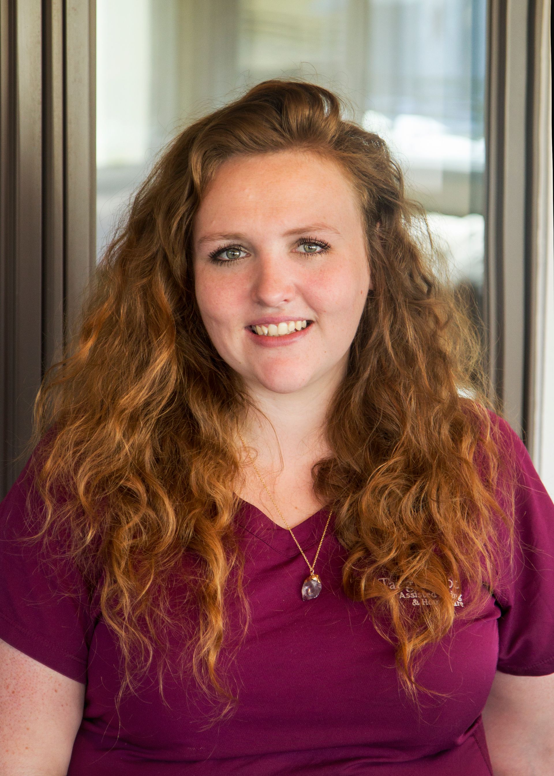 Woman with long, reddish-brown curly hair smiles in front of a window, wearing a dark red top and necklace.