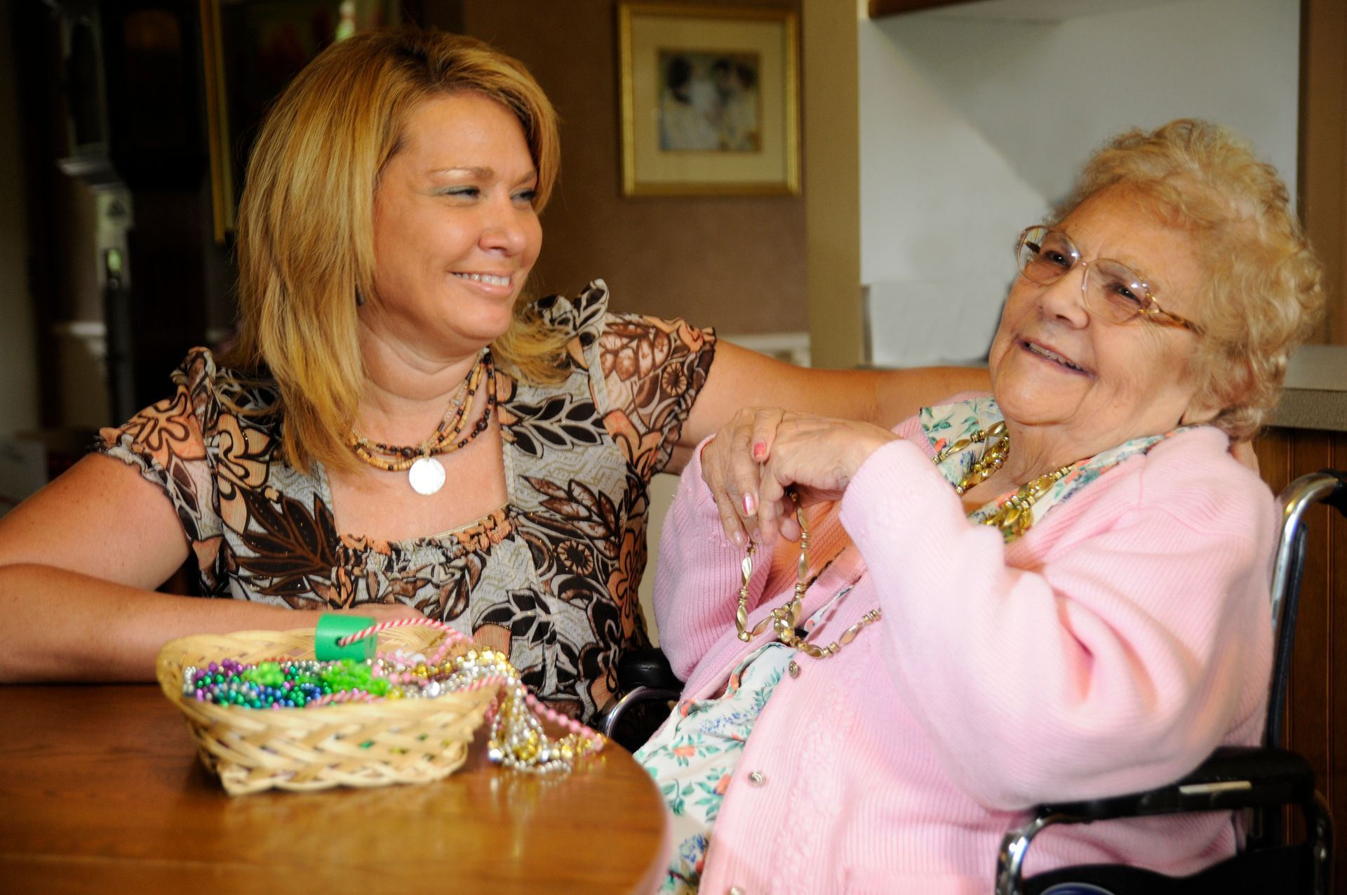 Woman smiles at an elderly person seated in a wheelchair; they are indoors and appear happy.