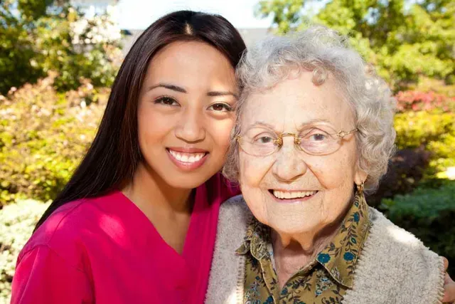 Woman in pink top smiles with elderly woman outdoors.