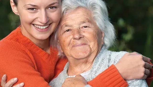 Woman in orange sweater hugs an elderly woman. Both smile, outside with green background.
