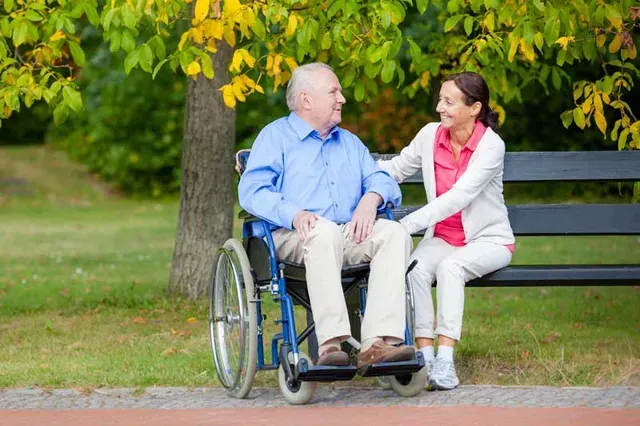 Man in wheelchair with caregiver on park bench, smiling. Tree with yellow leaves in the background.