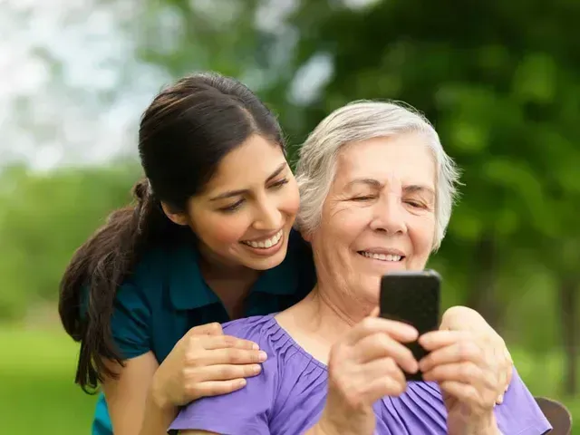 Woman helps older person use a smartphone outdoors; both smile.