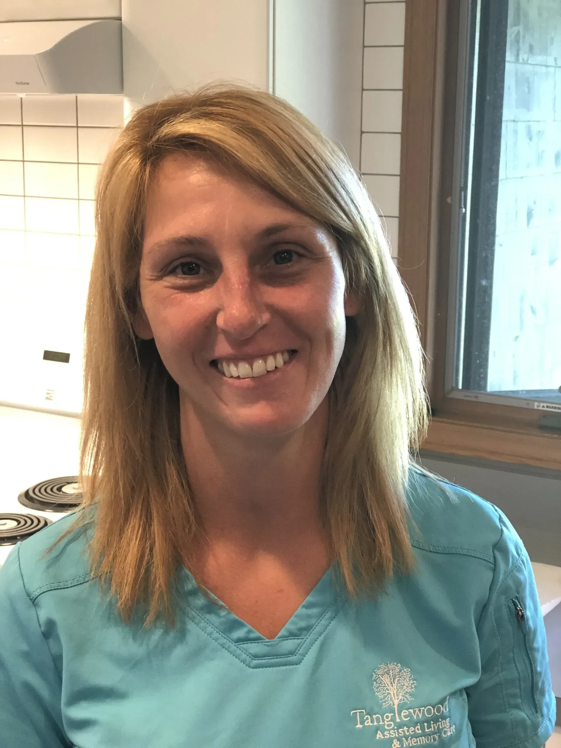 Woman with blonde hair wearing a blue scrub top, smiling. Standing near a window, kitchen visible.