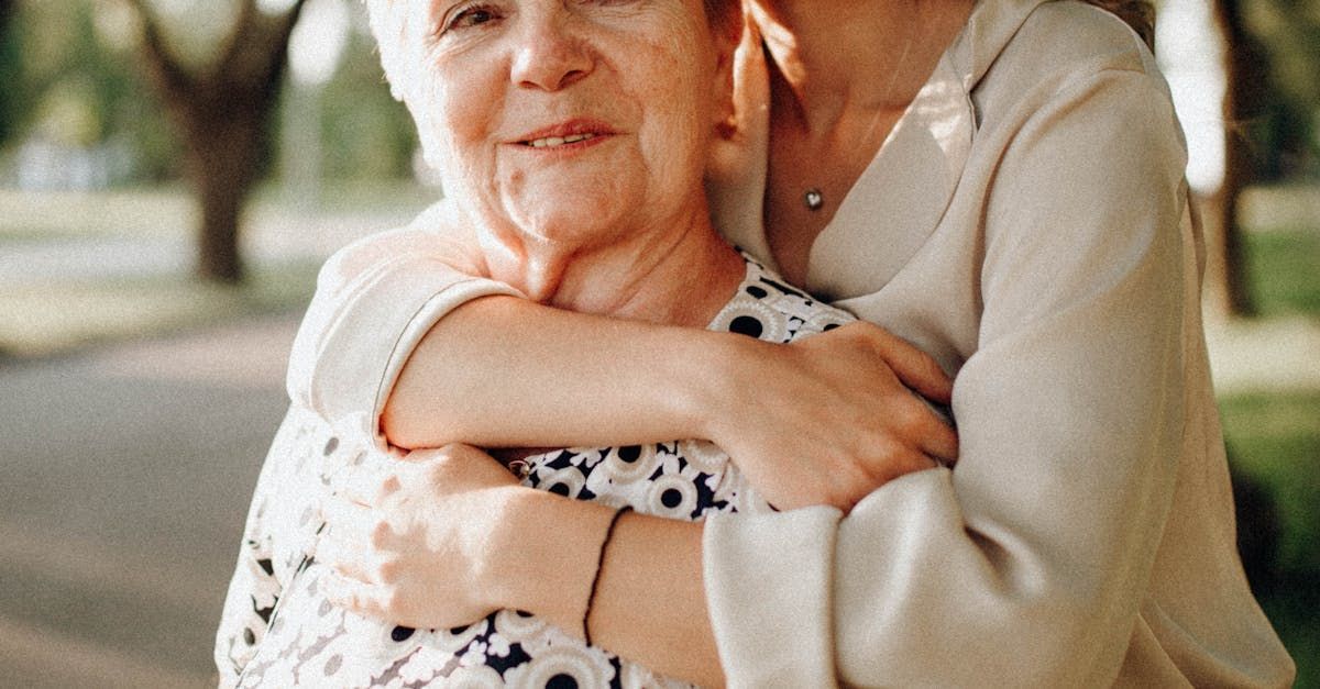 Woman embraces an older woman outdoors. Both wear light-colored tops, smiling. Sunlight and trees in background.