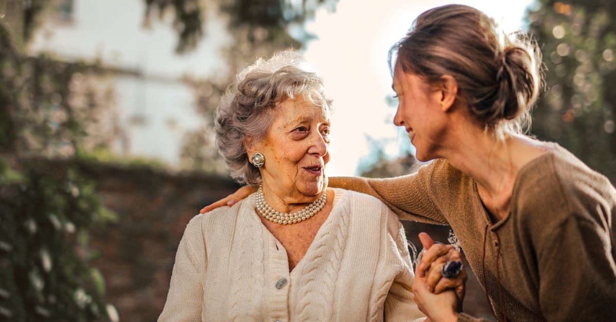 Woman with hand on elderly person's shoulder, smiling outdoors, soft sunlight.