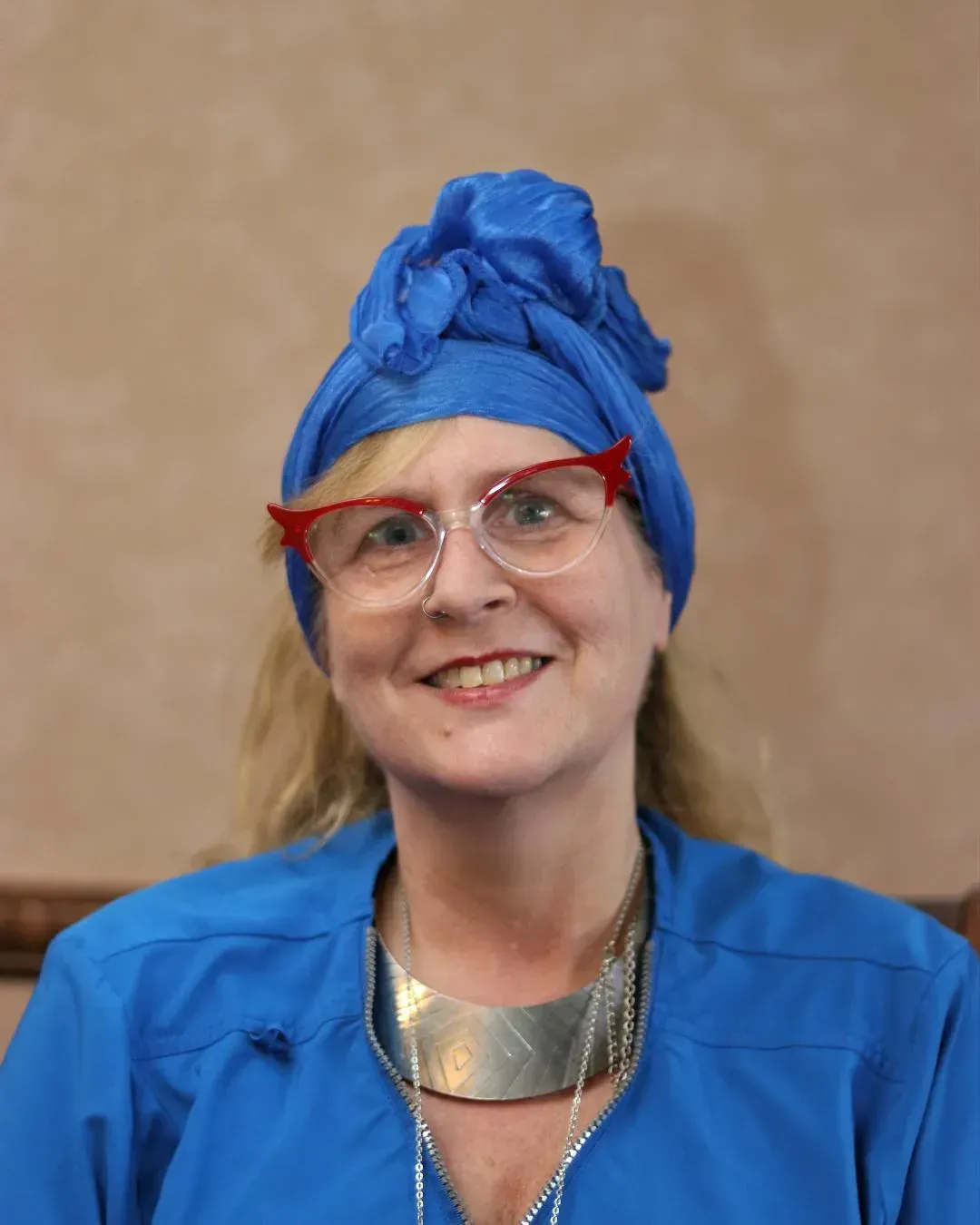 Woman with blue headscarf, glasses, and necklace, wearing a blue shirt, smiling.