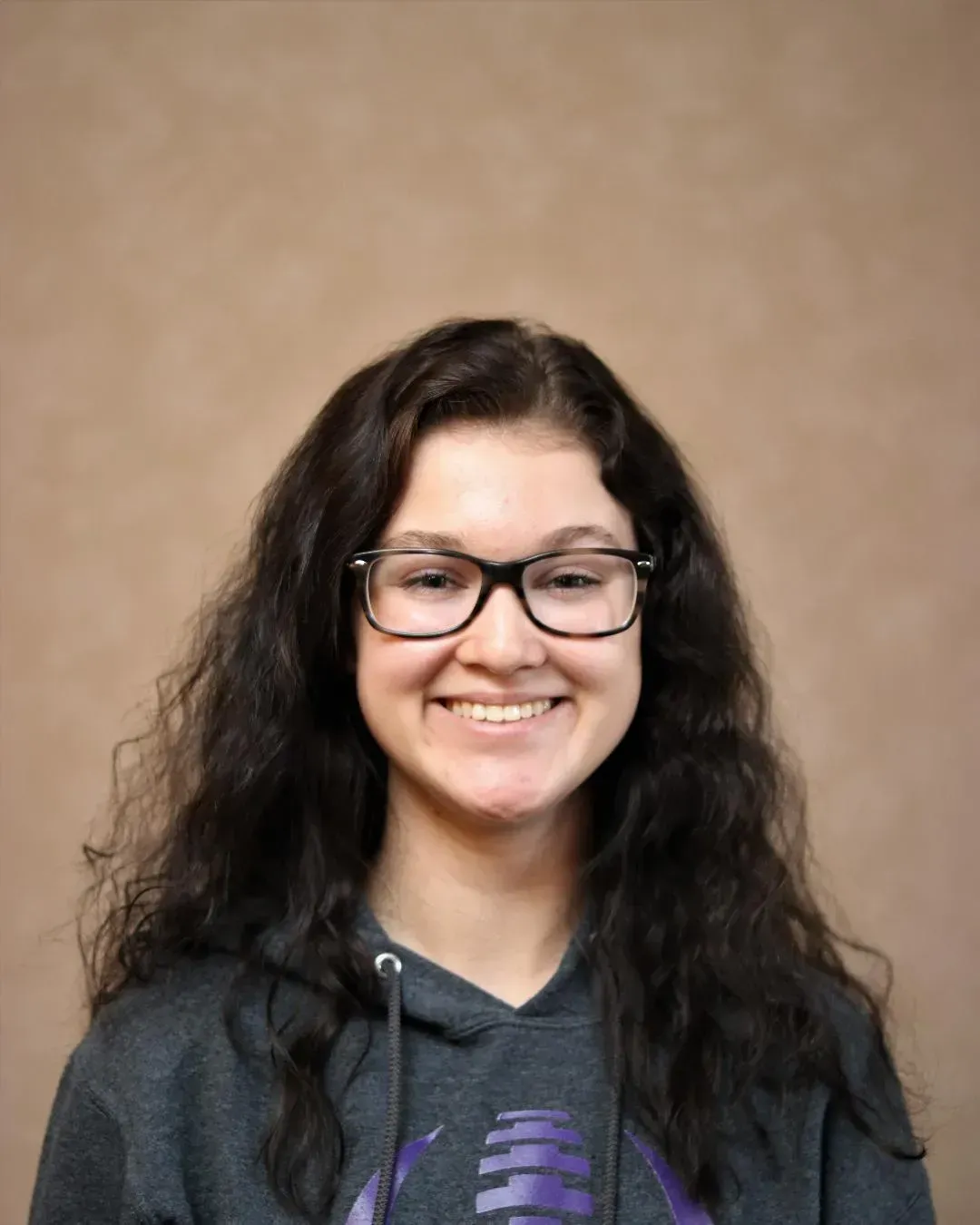 Woman with glasses smiles, wearing a gray hoodie, in front of a neutral backdrop.