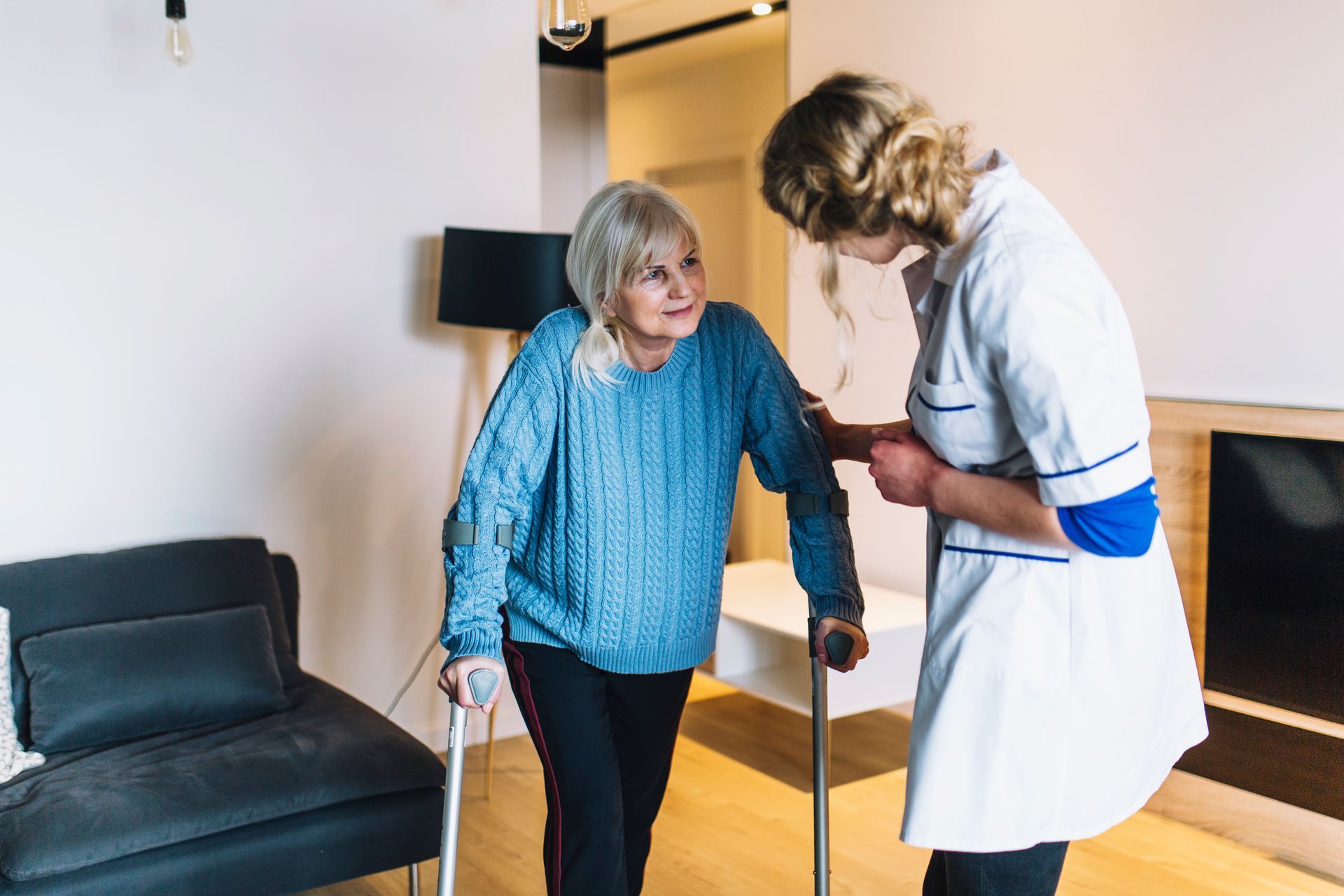 Woman using crutches assisted by healthcare worker in a home setting.