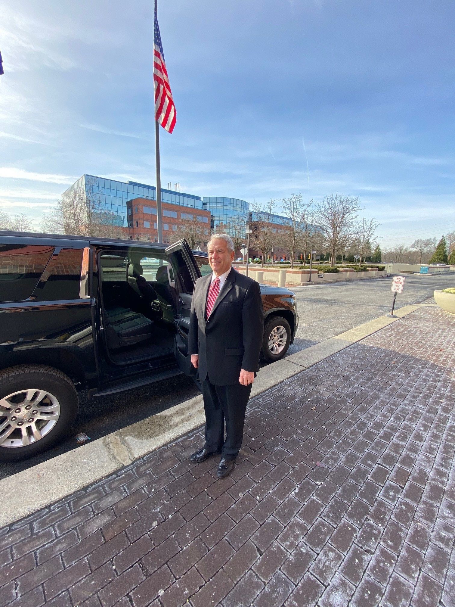Man in suit stands beside a black SUV with an American flag and a building in the background.
