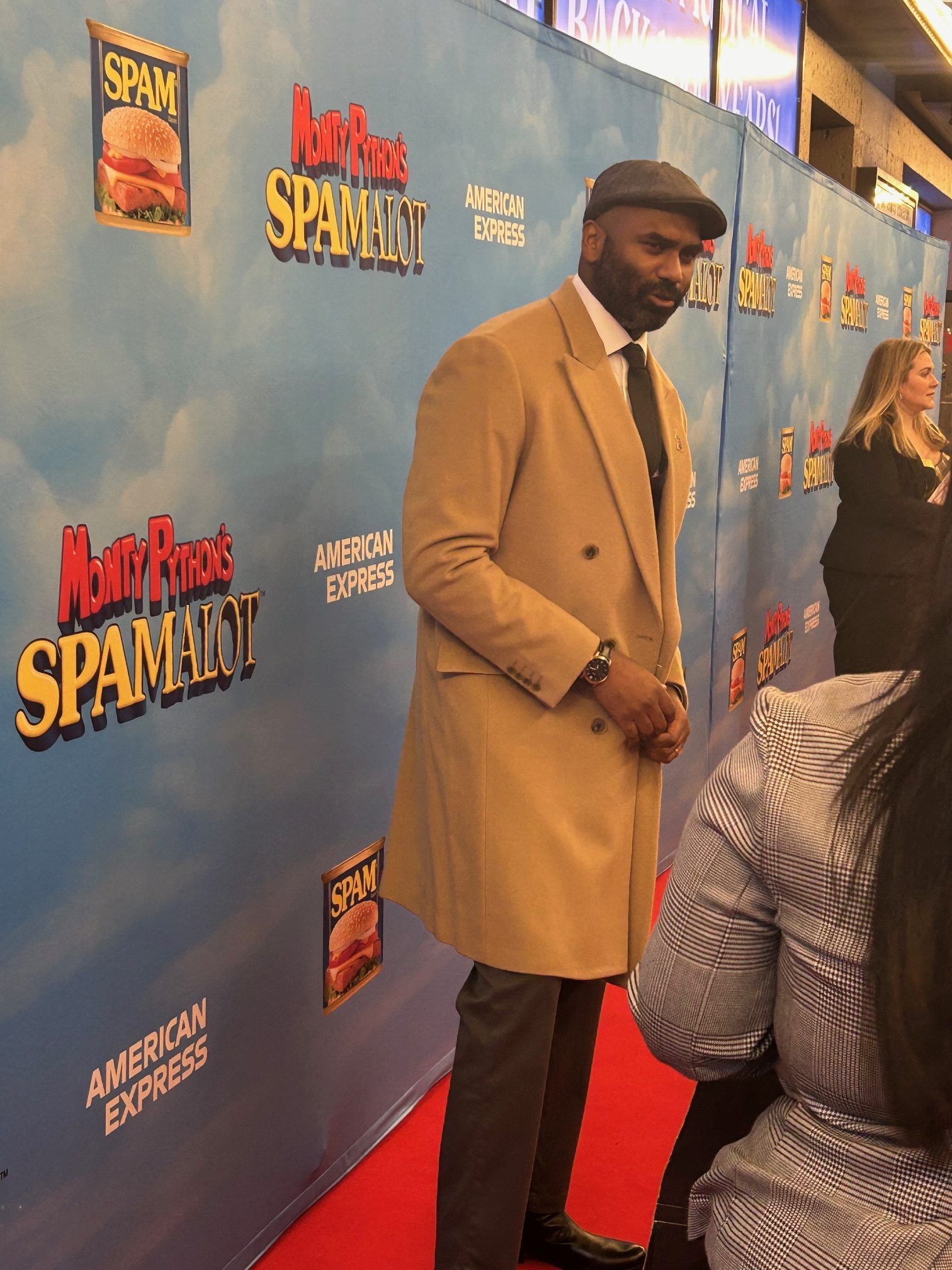 Man in tan coat and cap posing on a red carpet with a