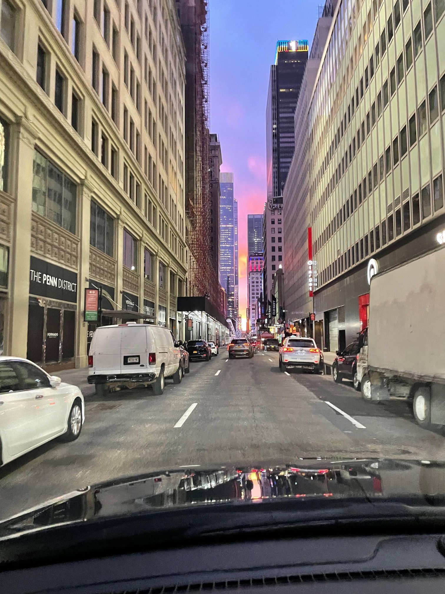 Street view of a city with tall buildings, cars, and a colorful sunset.