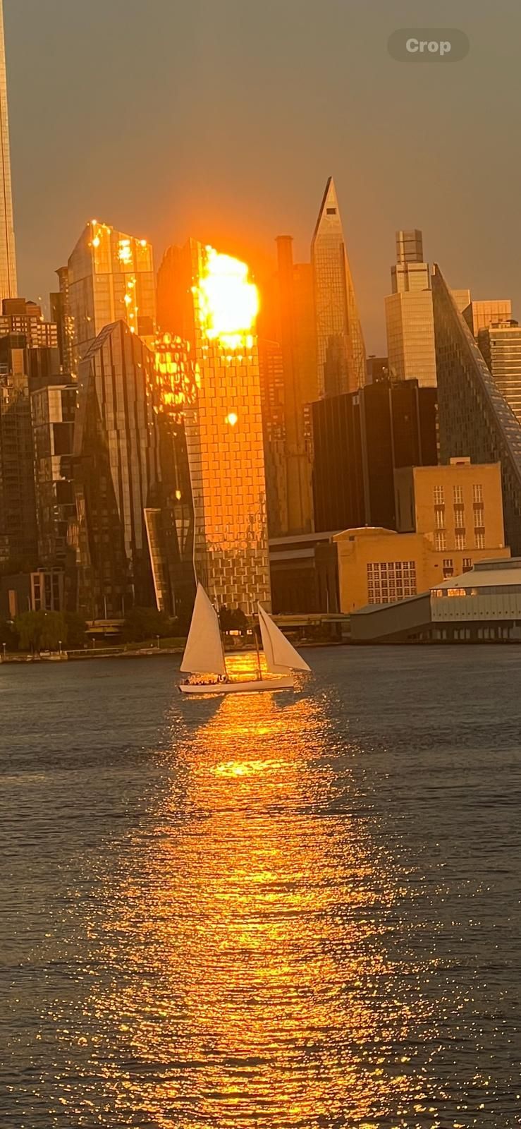 Sailboat on water reflecting golden sunlight, city skyline in background.