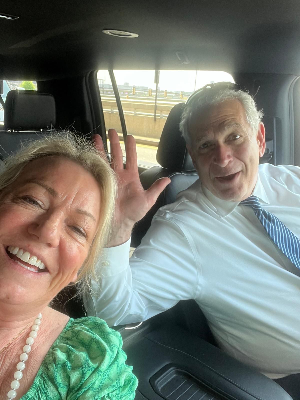 Woman and man smiling, waving from inside a car; woman in green, man in white shirt and tie.