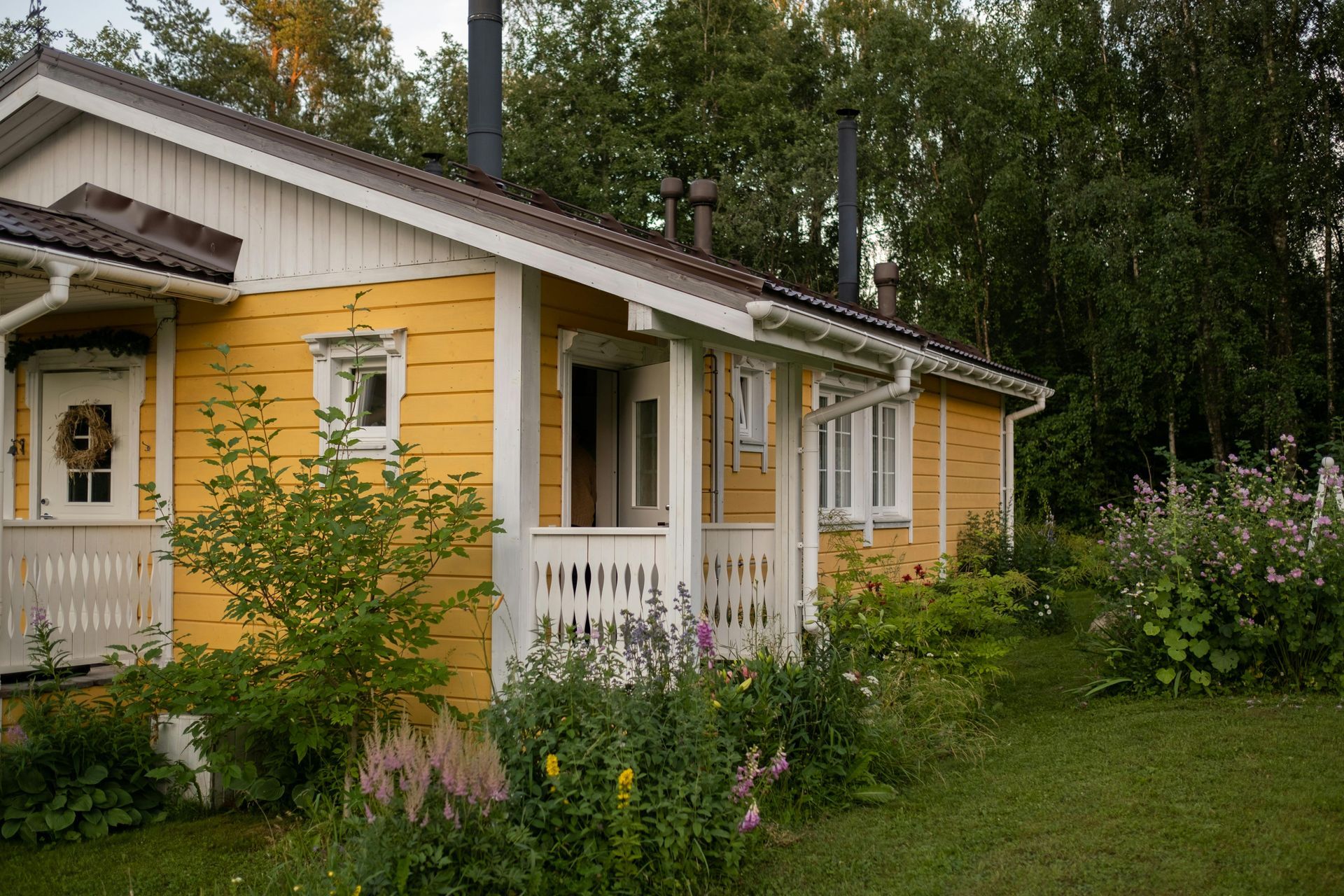 Yellow house with white trim, porch, and garden. Green trees in the background.
