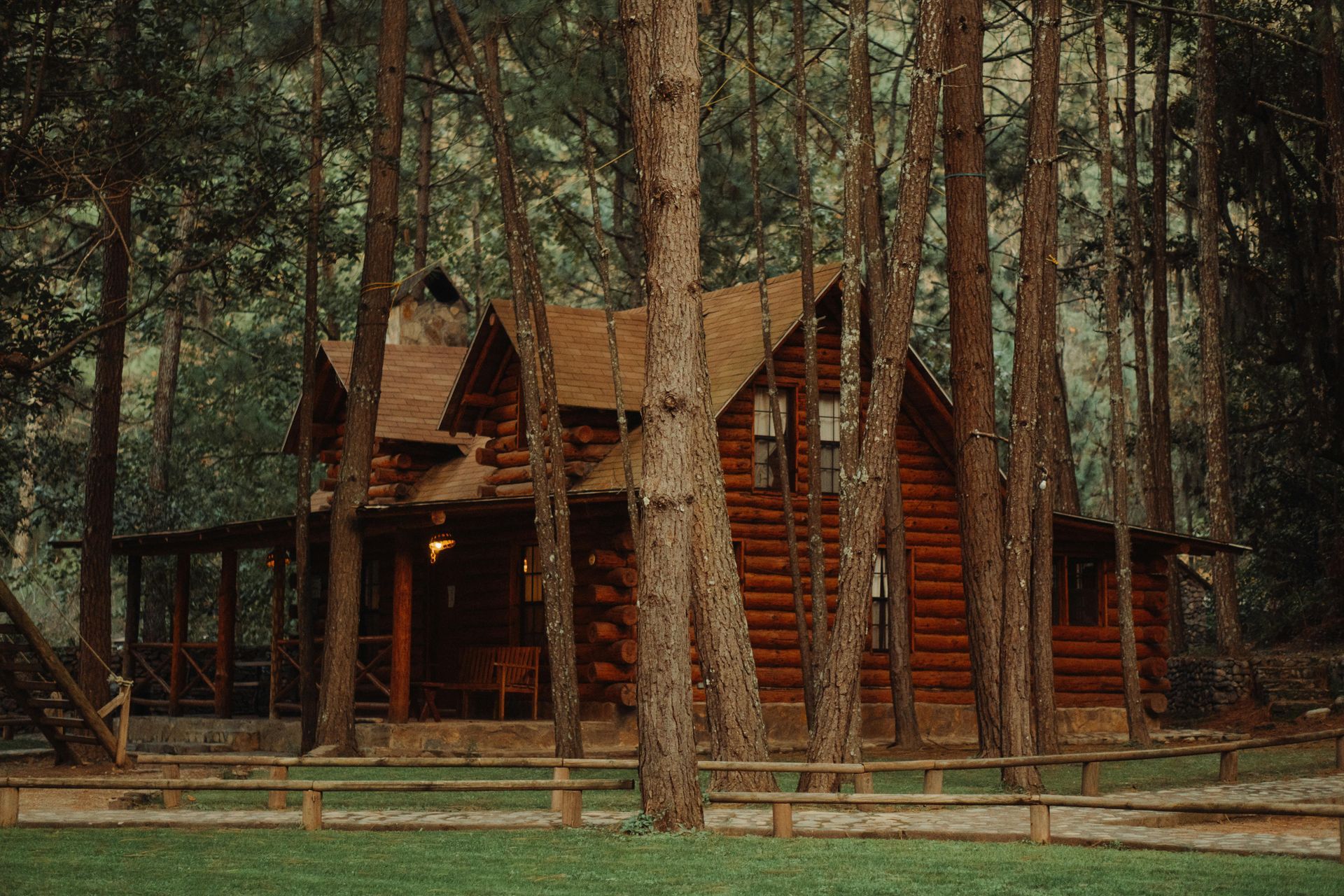 Log cabin nestled in a forest, brown logs, wooden porch, surrounded by trees and a low fence.