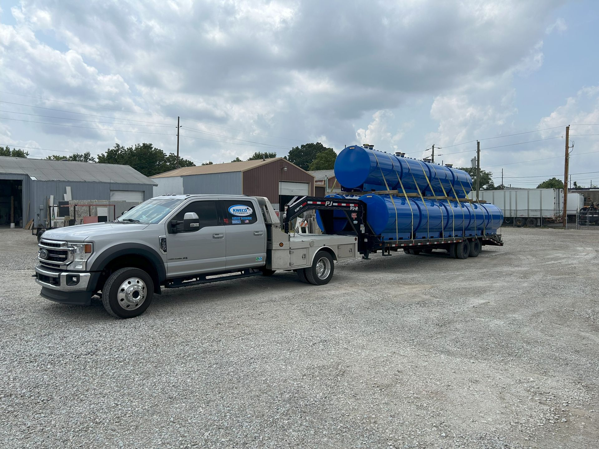 A silver Ford truck towing a trailer with three large blue cylindrical tanks in a gravel lot on a cloudy day.