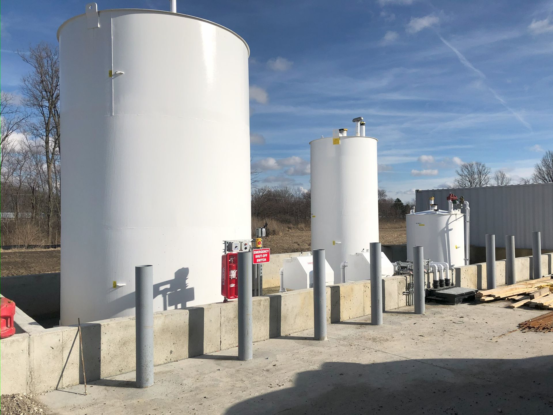 Three large white industrial tanks outdoors, next to concrete barriers and a gray fence, under a blue sky.
