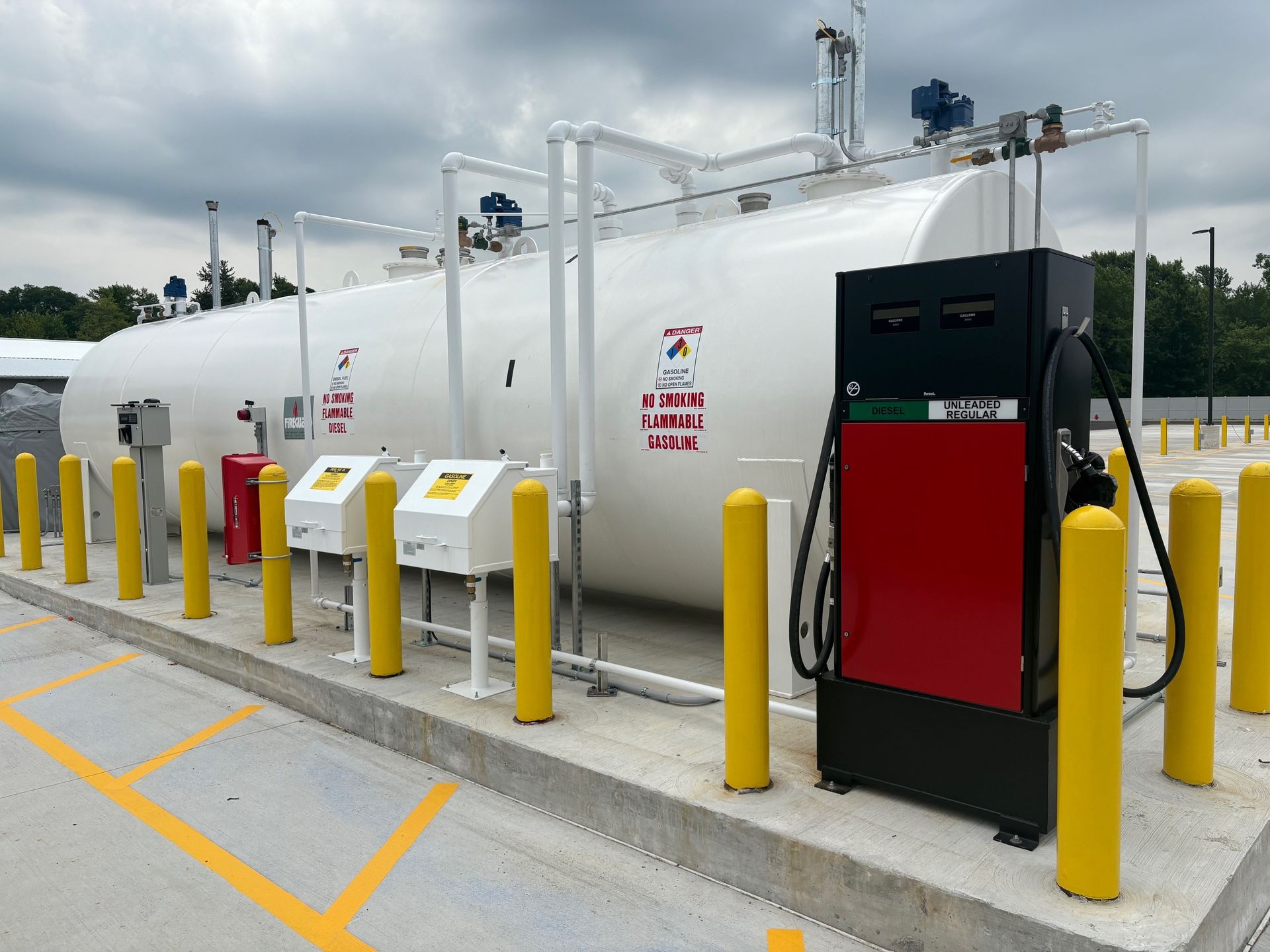 A white fuel tank and dispensing station, with a red pump, surrounded by yellow bollards on a concrete pad under a cloudy sky.