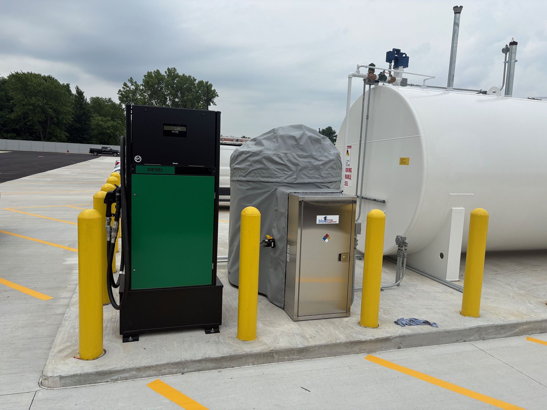 Fueling station with a large white tank, green and black machinery, and yellow bollards on a concrete pad.