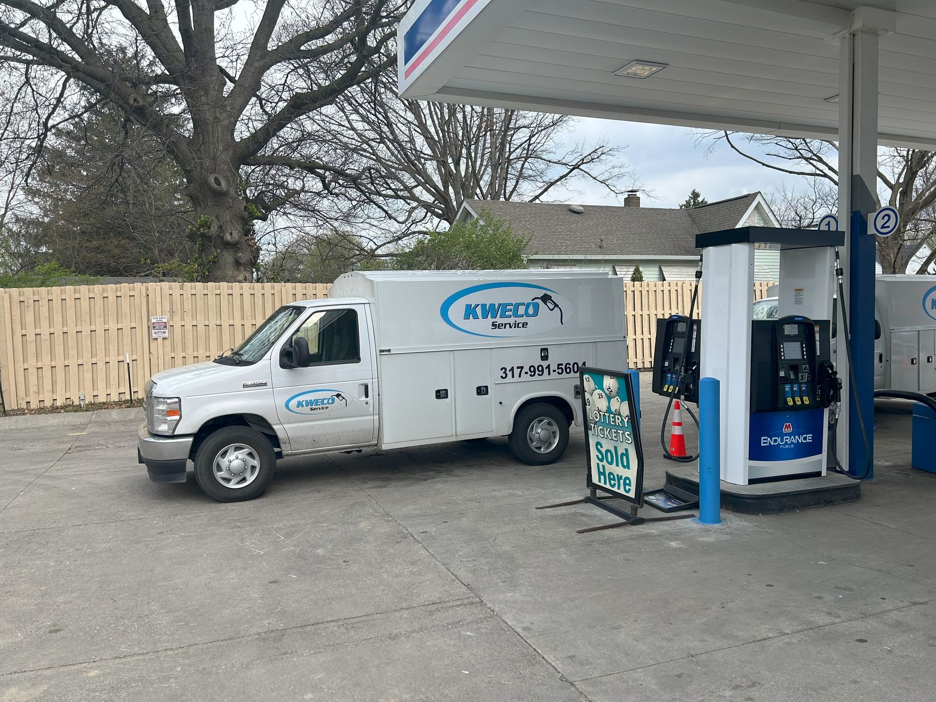 A white work van parked at a gas pump. The van has blue branding and a phone number. A fence and a house are in the background.