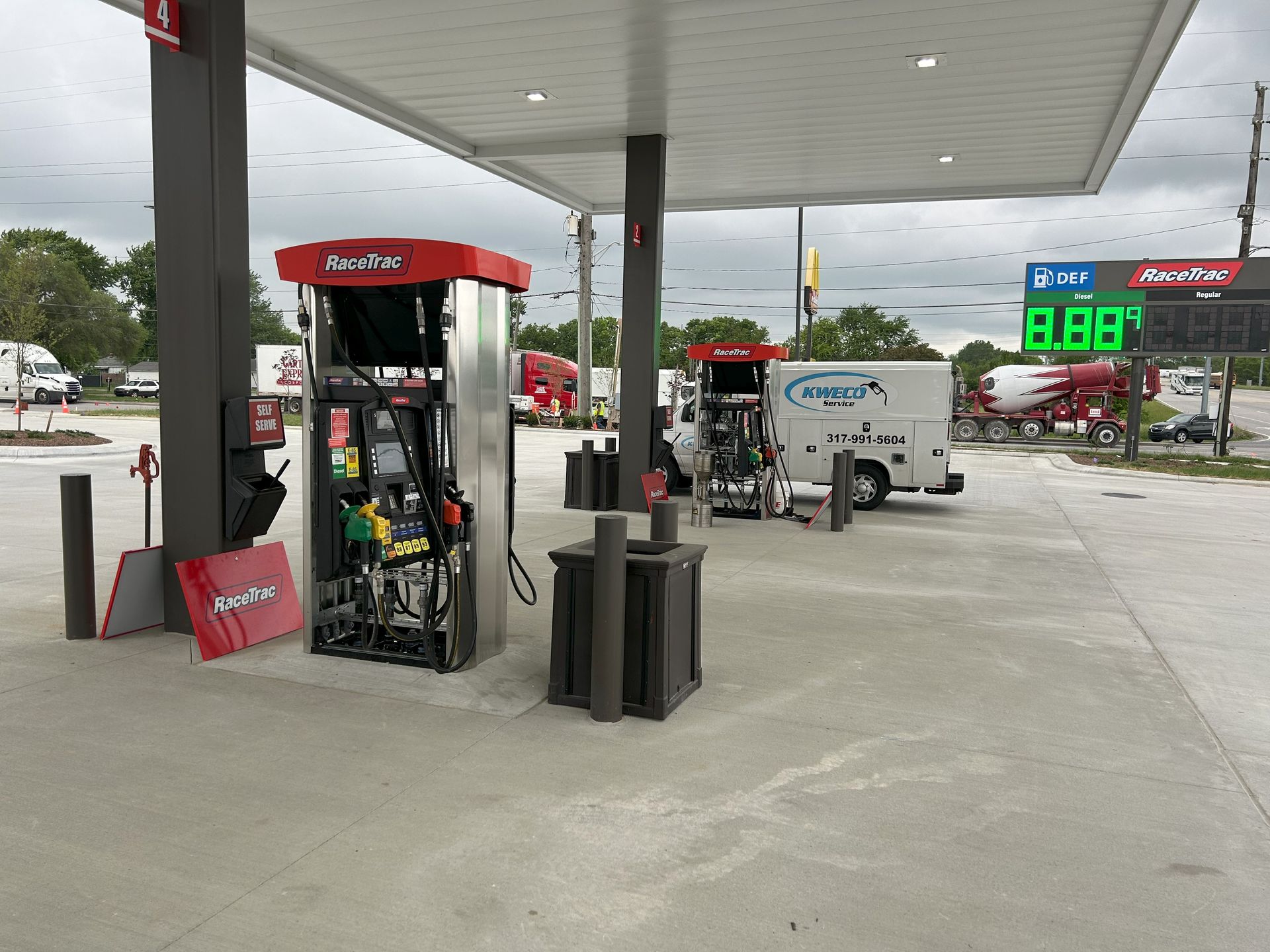 Gas station with red and gray fuel pumps under a canopy. A white service truck is parked nearby. The price of gas is displayed.