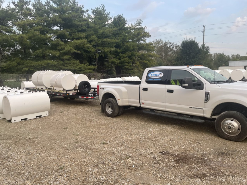 A white pickup truck towing a trailer loaded with white cylindrical tanks, likely for fuel storage, in an outdoor storage yard.
