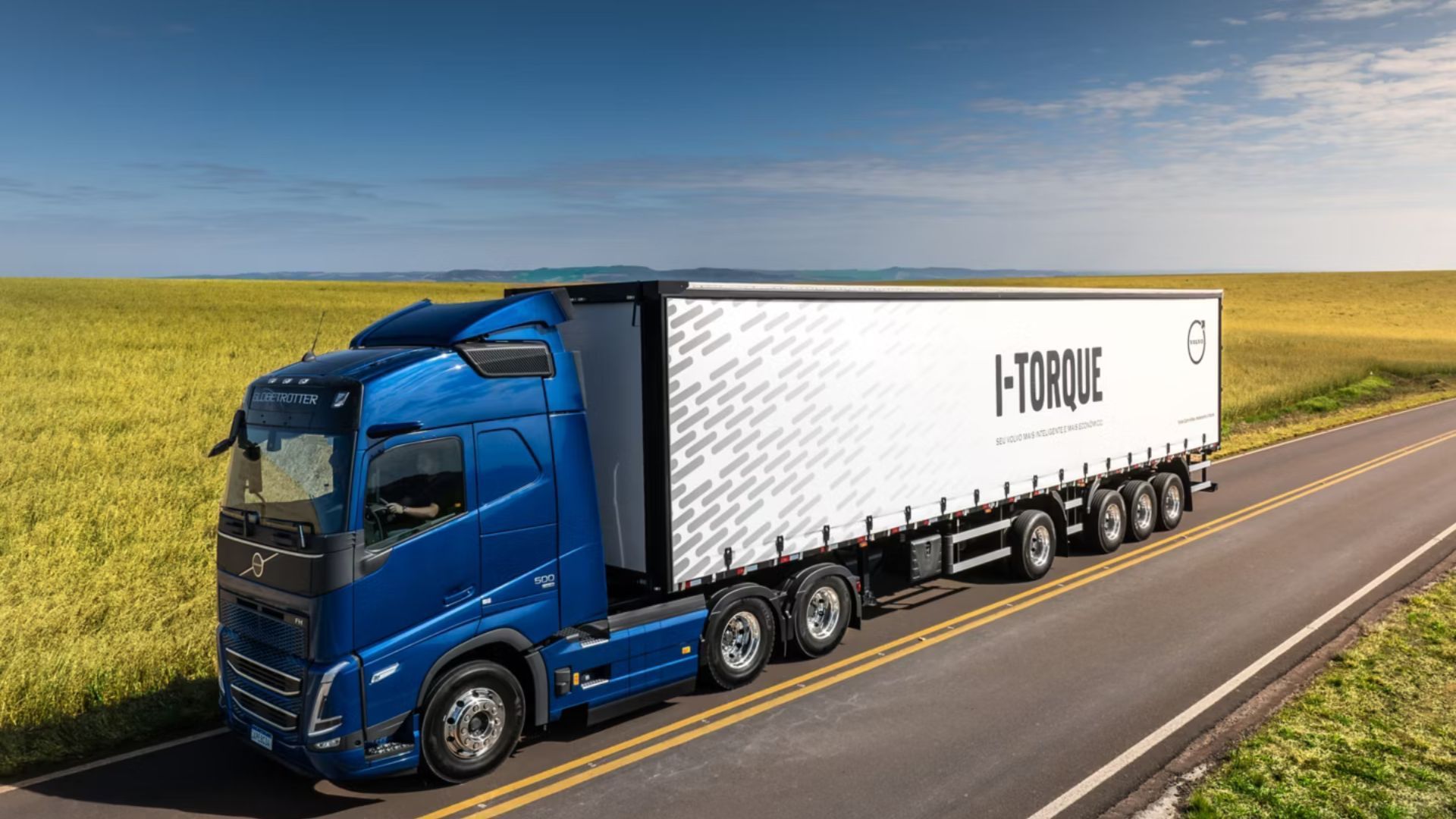 Blue semi-truck hauling a white trailer on a rural highway beside green fields